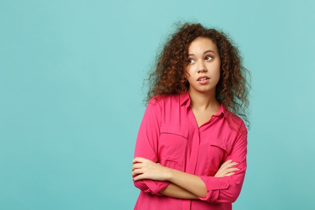 Ritratto di ragazza africana pensierosa in abiti casual rosa che guarda da parte, tenendo le mani incrociate isolate su sfondo blu turchese parete. persone sincere emozioni, concetto di stile di vita. mock up copia spazio.