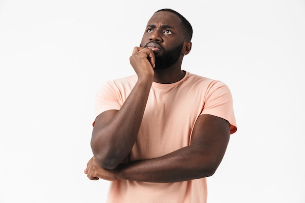 Portrait of a pensive african man wearing t-shirt standing isolated over white wall, thinking