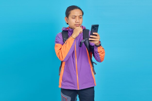 Portrait of pensive Asian young man looking at cell phone on blue background