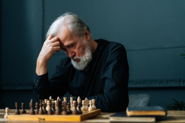 Portrait of pensive bearded mature adult pensioner male thinking about chess move sitting at home and playing chess alone