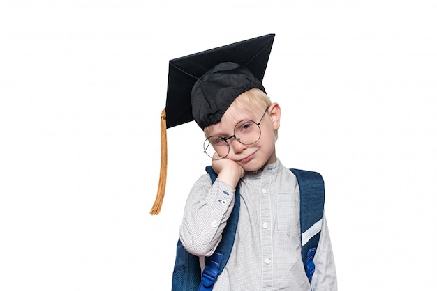 Portrait of a pensive blond boy in big glasses, an academic hat and a school bag. White background. Isolate