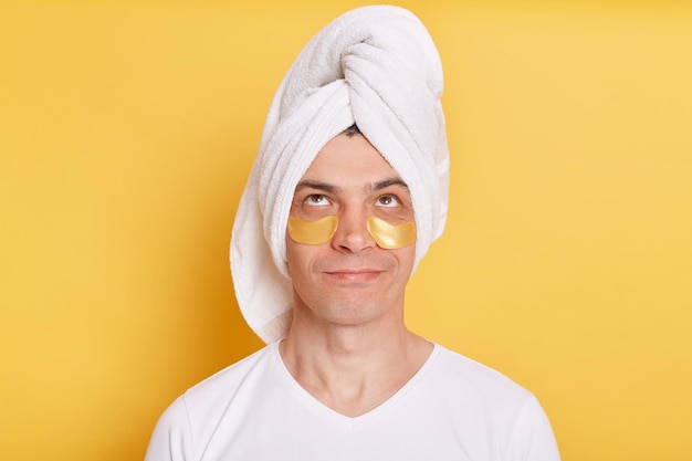 Portrait of pensive confused man wearing white t shirt and towel having cosmetic patches under his eyes looking up with puzzled facial expression posing isolated over yellow background