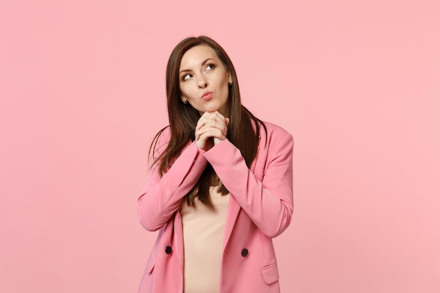 Portrait of pensive cute young woman in jacket looking up, put hands prop up on chin isolated on pastel pink wall background in studio. People sincere emotions, lifestyle concept. Mock up copy space.