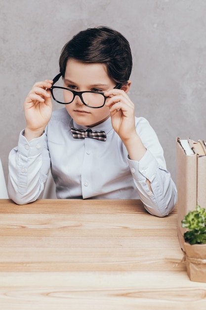 Portrait of pensive little boy in eyeglasses sitting at workplace isolated on grey