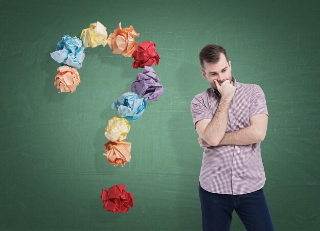 Portrait of a pensive man wearing a pink shirt and standing near a green chalkboard with a question mark made of colored paper balls