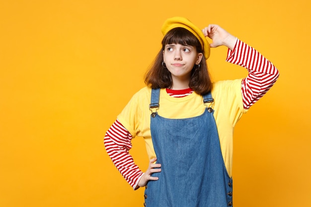 フランスのベレー帽、頭に手を置いて、黄色の背景に孤立して見上げるデニムのサンドレスで物思いにふける困惑した少女ティーンエイジャーの肖像画。人々の誠実な感情のライフスタイルの概念。コピースペースをモックアップします。