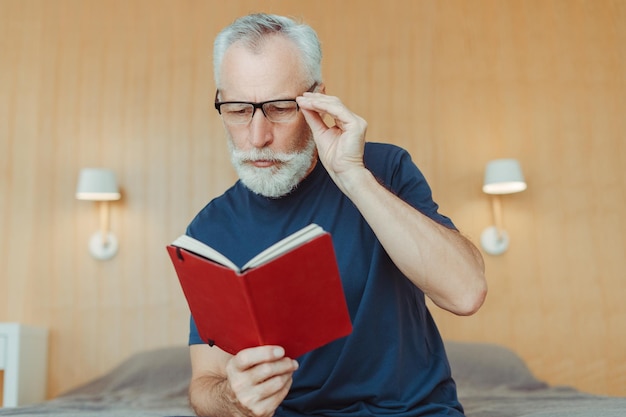 Portrait of pensive senior man wearing glasses holding notebook reading book sitting on bed in living room Elderly pensioner in casual clothes studying Education concept