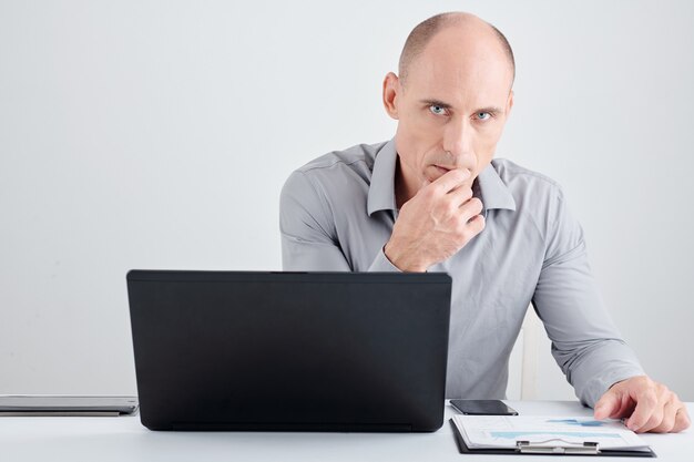 Portrait of pensive serious entrepreneur sitting at office desk with laptop and report and looking at camera