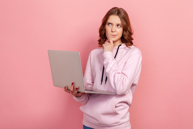 Ritratto di una ragazza adolescente dai capelli ricci e intelligente con felpa con cappuccio che tiene un laptop e pensa al progetto scolastico scatto in studio al coperto isolato su sfondo rosa