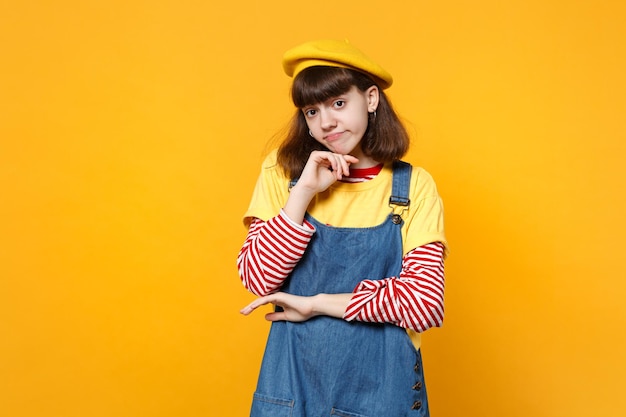 Portrait of pensive upset girl teenager in french beret, denim sundress put hand prop up on chin isolated on yellow wall background. people sincere emotions, lifestyle concept. mock up copy space