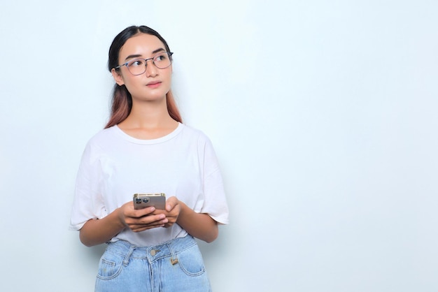 Portrait of pensive young Asian girl in white tshirt holding smartphone looking away at copy space isolated on white background