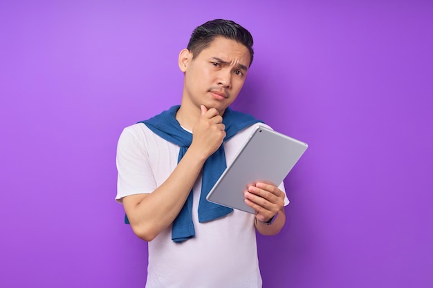 Portrait of pensive young Asian man in white tshirt touching chin and holding digital tablet isolated on purple background