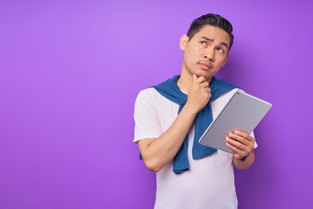 Portrait of pensive young Asian man in white tshirt touching chin and holding digital tablet isolated on purple background