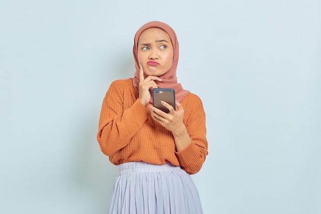 Portrait of pensive young asian muslim woman in brown sweater\
hijab holding mobile phone isolated on white background muslim\
lifestyle concept