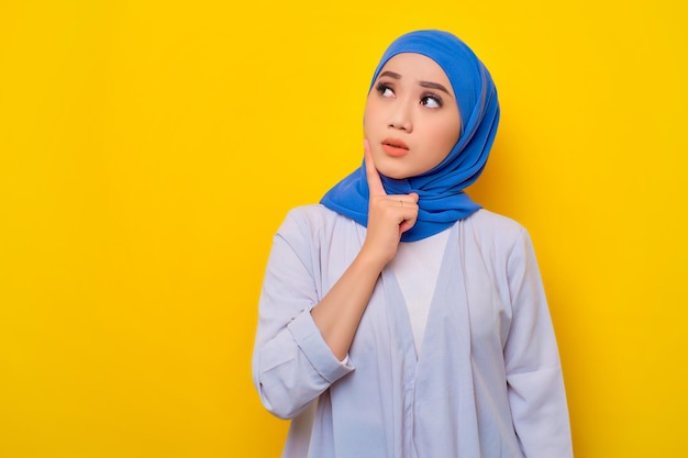 Portrait of pensive young asian muslim woman looking up at copy
space thinking about something isolated over yellow background