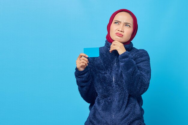 Portrait of pensive young asian woman holding credit card and looking up on blue background