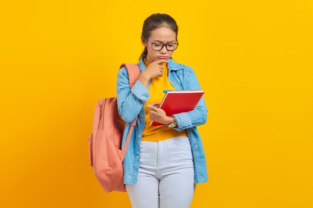 책을 들고 배낭을 메고 캐주얼 옷을 입은 수심에 찬 젊은 아시아 여성 학생의 초상화 대학 대학 개념의 노란색 배경 교육에 대해 생각하는 질문