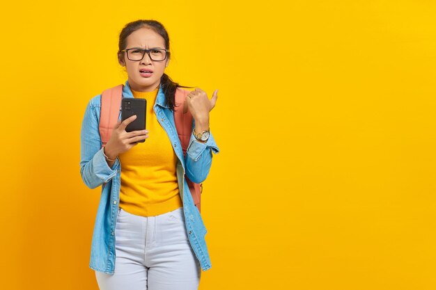 Portrait of pensive young Asian woman student in casual clothes with backpack using mobile phone and pointing at copy space with fingers isolated on yellow background