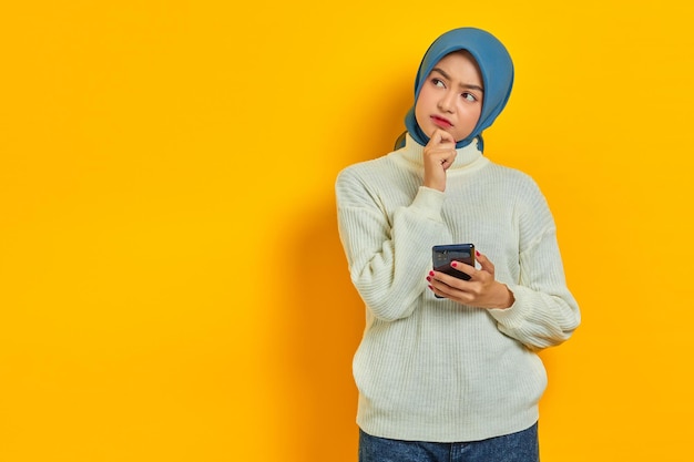 Portrait of pensive young asian woman in white sweater and\
hijab thinking about question with hand on chin while holding\
mobile phone isolated over yellow background people islam religious\
concept