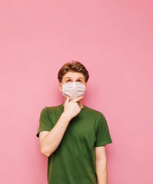 분홍색 배경에 격리된 녹색 티셔츠와 흰색 의료용 마스크를 쓴 수심에 찬 젊은이의 초상화