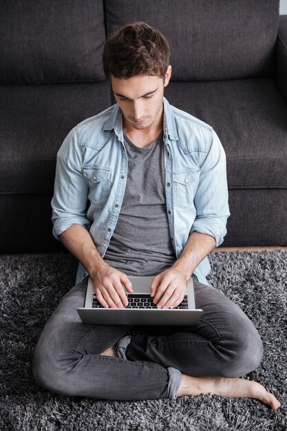 Portrait of a pensive young man typing on laptop computer while sitting on carpet at home