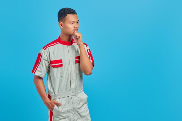 Portrait of pensive young mechanic touching chin with hand and looking sideways on blue background