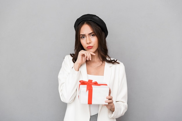 Portrait of a pensive young woman dressed in jacket over gray background, holding present box