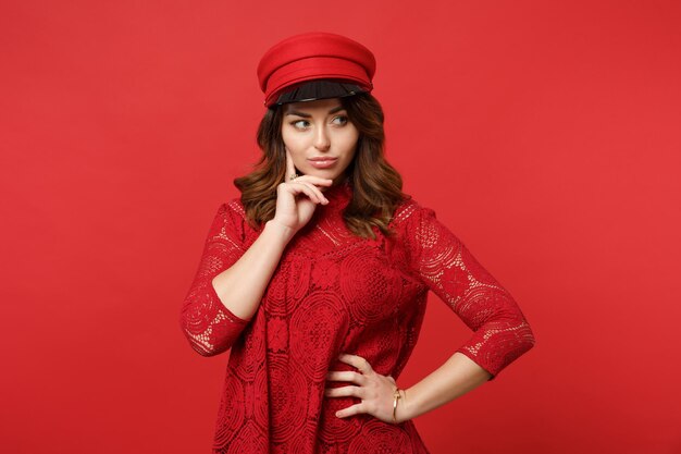 Portrait of pensive young woman in lace dress, cap looking aside, put hand prop up on chin isolated on bright red background in studio. People sincere emotions, lifestyle concept. Mock up copy space.