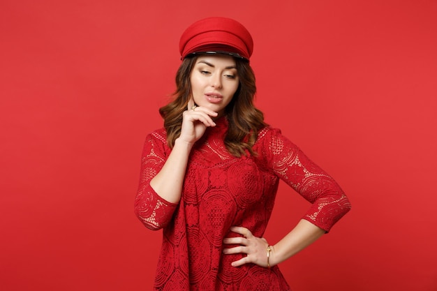 Portrait of pensive young woman in lace dress, cap looking down, put hand prop up on chin isolated on bright red background in studio. People sincere emotions, lifestyle concept. Mock up copy space.