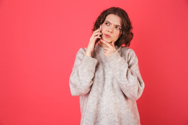 Portrait of a pensive young woman standing isolated over pink, talking on mobile phone