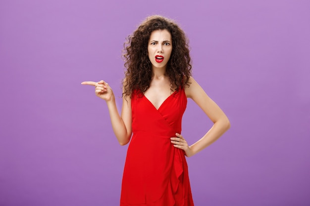 Portrait of perplexed and confused attractive curlyhaired female in red elegant dress standing perpl.