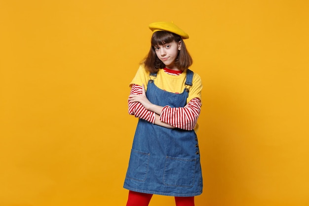 Portrait of perplexed girl teenager in french beret, denim sundress holding hands crossed isolated on yellow wall background in studio. People sincere emotions, lifestyle concept. Mock up copy space.