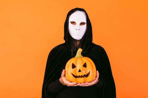 Portrait of a person with white Halloween killer mask, cape and hood, holding a pumpkin jack-o'-lantern, dressed for Halloween over orange background