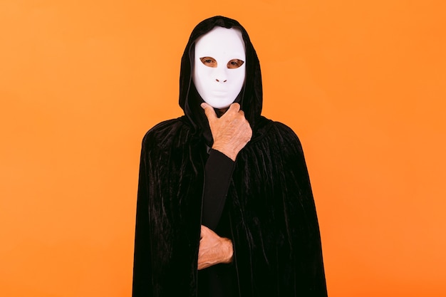 Portrait of person with white Halloween killer mask, cape and hood looking at camera, touching chin with hand, dressed for Halloween over orange background