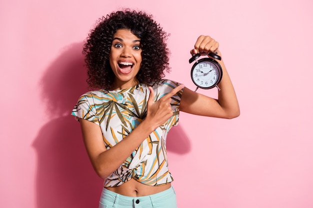 Foto ritratto di una donna africana riccia che indica l'orologio sorridendo allegramente indossando abiti casual isolati su sfondo di colore rosa pastello