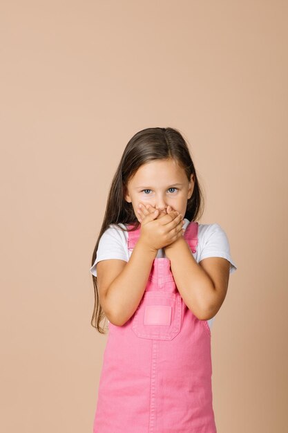 Portrait photo of female kid closing mouth with hands not talk with others with shining eyes and sullen look looking at camera wearing bright pink jumpsuit and white tshirt on beige background
