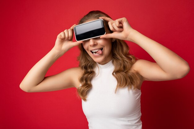 Портретное фото смешной молодой блондинки в белой футболке, изолированной на красном фоне с копией пространства, держащей смартфон, показывающий телефон в руке с пустым экраном для выреза и показывающий язык.