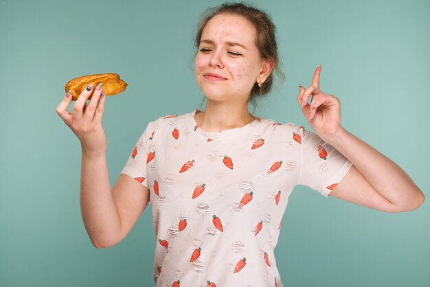 Il ritratto della ragazza teenager brufolosa vuole mangiare la torta di eclair sull'azzurro