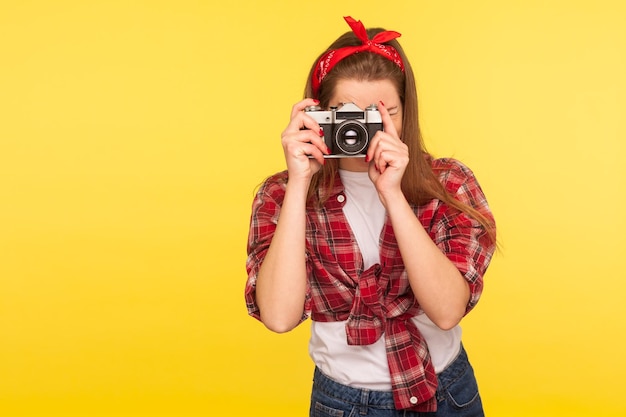 チェッカーシャツとヘッドバンドのピンナップガールの肖像画は、昔ながらのカメラ旅行者と一緒に写真を撮って、黄色の背景に分離されたレトロな5039年代スタイルのスタジオショットを楽しんで写真を撮る