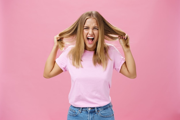 Ritratto di donna arrabbiata e indignata che si sente sotto pressione, urla e si strappa i capelli dalla testa in piedi infastidita su sfondo rosa