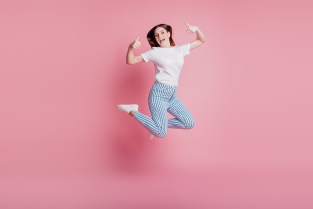 Ritratto di giocosa ragazza pazza che salta in aria alza due pollici su sfondo rosa