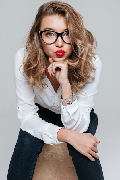 Portrait of a playful funny woman in sunglasses sitting on chair  isolated on a white wall