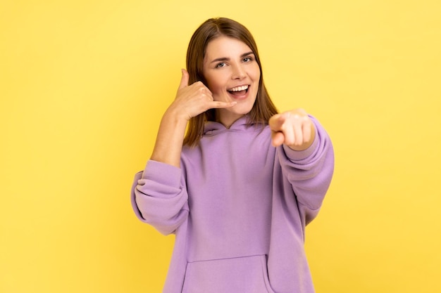 Portrait of playful happy woman making telephone gesture and pointing to camera