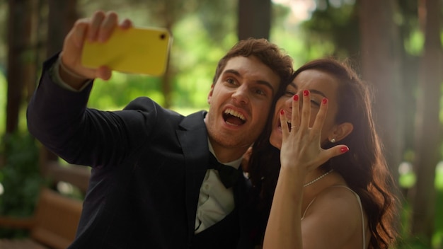 Photo portrait of playful man and woman doing selfie photo on mobile phone outdoors closeup joyful wedding couple making hand gestures in park handsome groom doing facial expression in garden