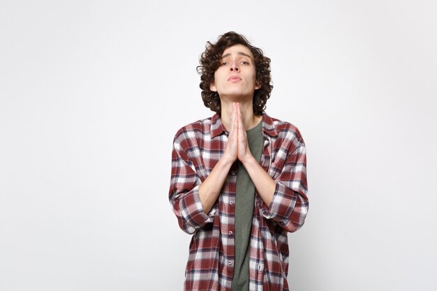 Portrait of pleading young man in casual clothes looking camera, folded his hands, praying isolated on white wall background in studio. People sincere emotions, lifestyle concept. Mock up copy space.