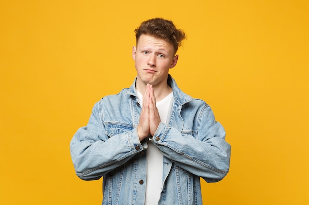 Portrait of pleading young man in denim casual clothes looking camera praying, holding hands folded isolated on yellow orange background. People sincere emotions lifestyle concept. Mock up copy space.