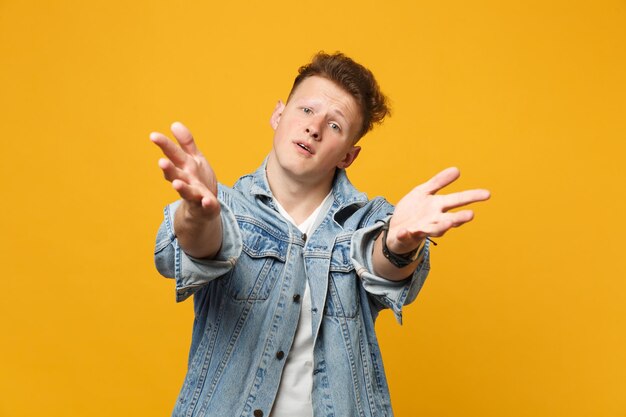 Portrait of pleading young man in denim casual clothes standing with outstretching hands isolated on yellow orange background in studio. people sincere emotions lifestyle concept. mock up copy space