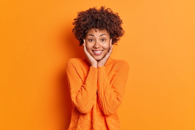 Portrait of pleasant looking woman keeps hands on face admires something smiles gently and looks directly at camera wears sweater isolated over vivid orange wall