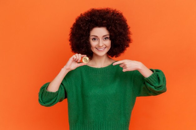 Портрет приятно выглядящей женщины с афро-прической в зеленом свитере в стиле кэжуал, указывающей пальцем на золотой биткойн, электронную коммерцию. Крытая студия снята на оранжевом фоне.