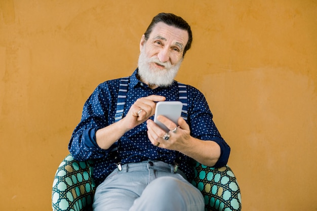 Portrait of pleasant senior smiling bearded man, wearing stylish shirt and pants, sitting on the yellow background and using the phone 